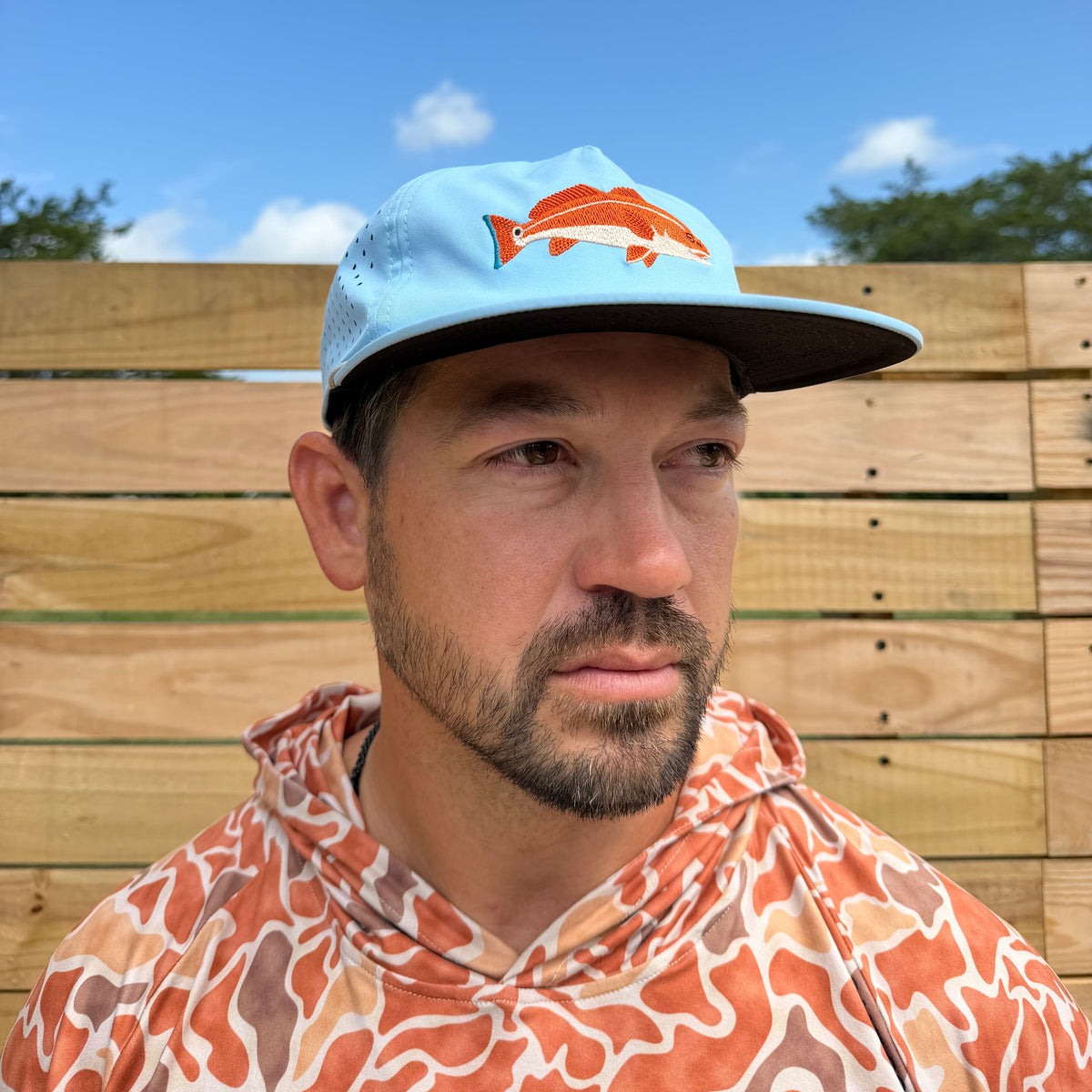 A man wearing the Salt SQD Redfish Cap  paired with an orange and white patterned shirt, photographed outdoors against a wooden fence and blue sky.