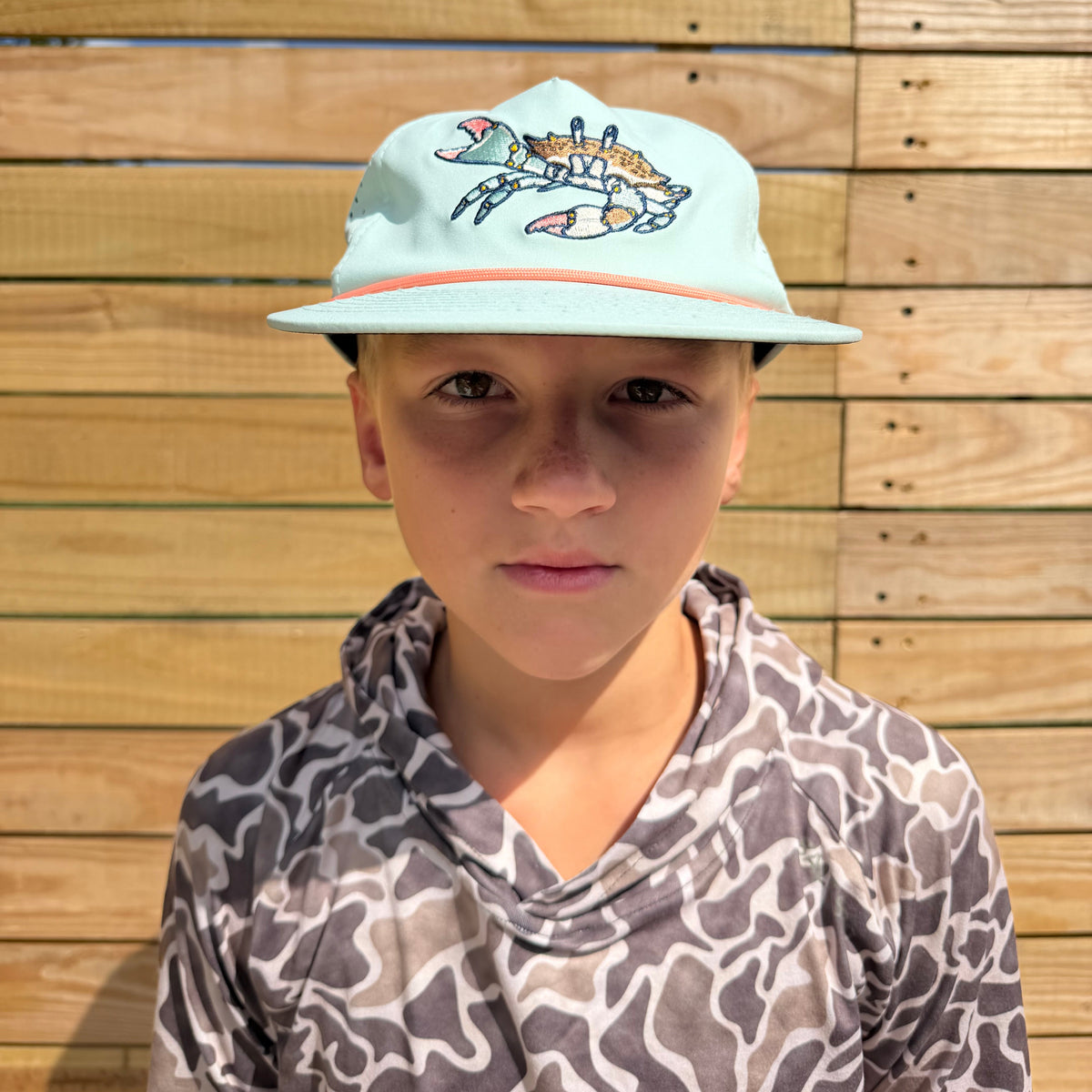 A boy wearing a Salt SQD Coastal Icons Cap in Blue Crab, photographed from the front, showing the crab logo and rope brim, paired with a camo long-sleeve shirt against a wooden plank background