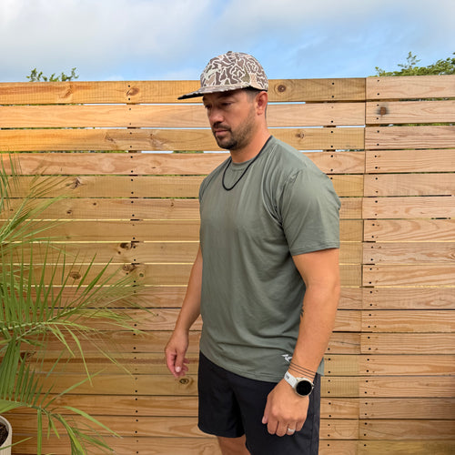 SqdDrift Salt SQD tee in Marsh Green with four-way stretch fabric, shown outdoors against a wooden fence.