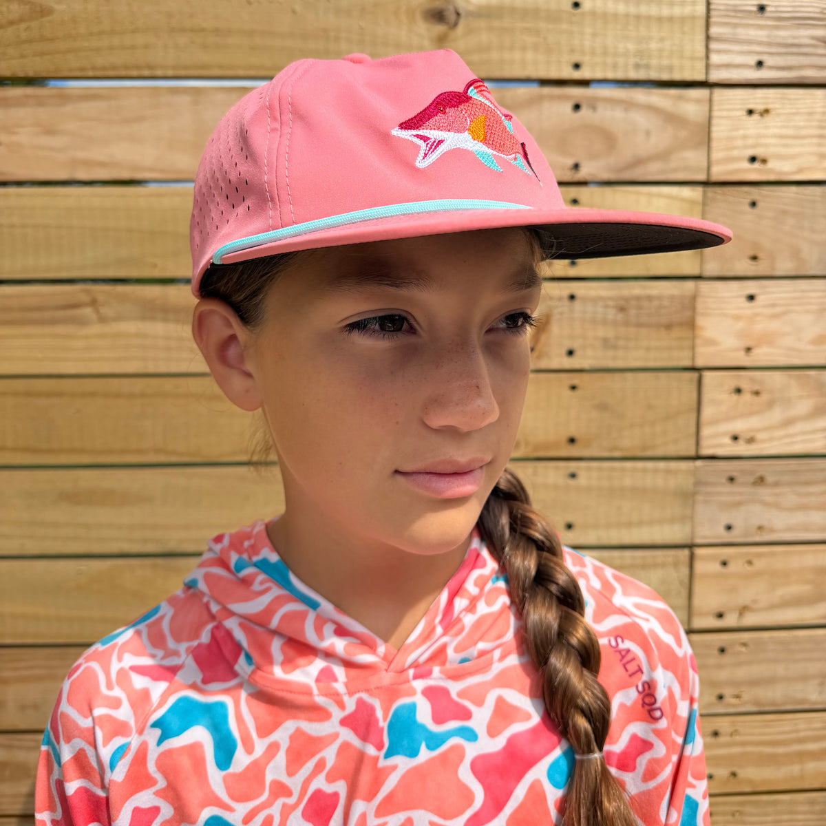 A young girl with a braided ponytail wearing a Salt SQD Coastal Icons Cap in Hogfish, front view showing hogfish logo and rope brim, photographed against a light wood plank wall.