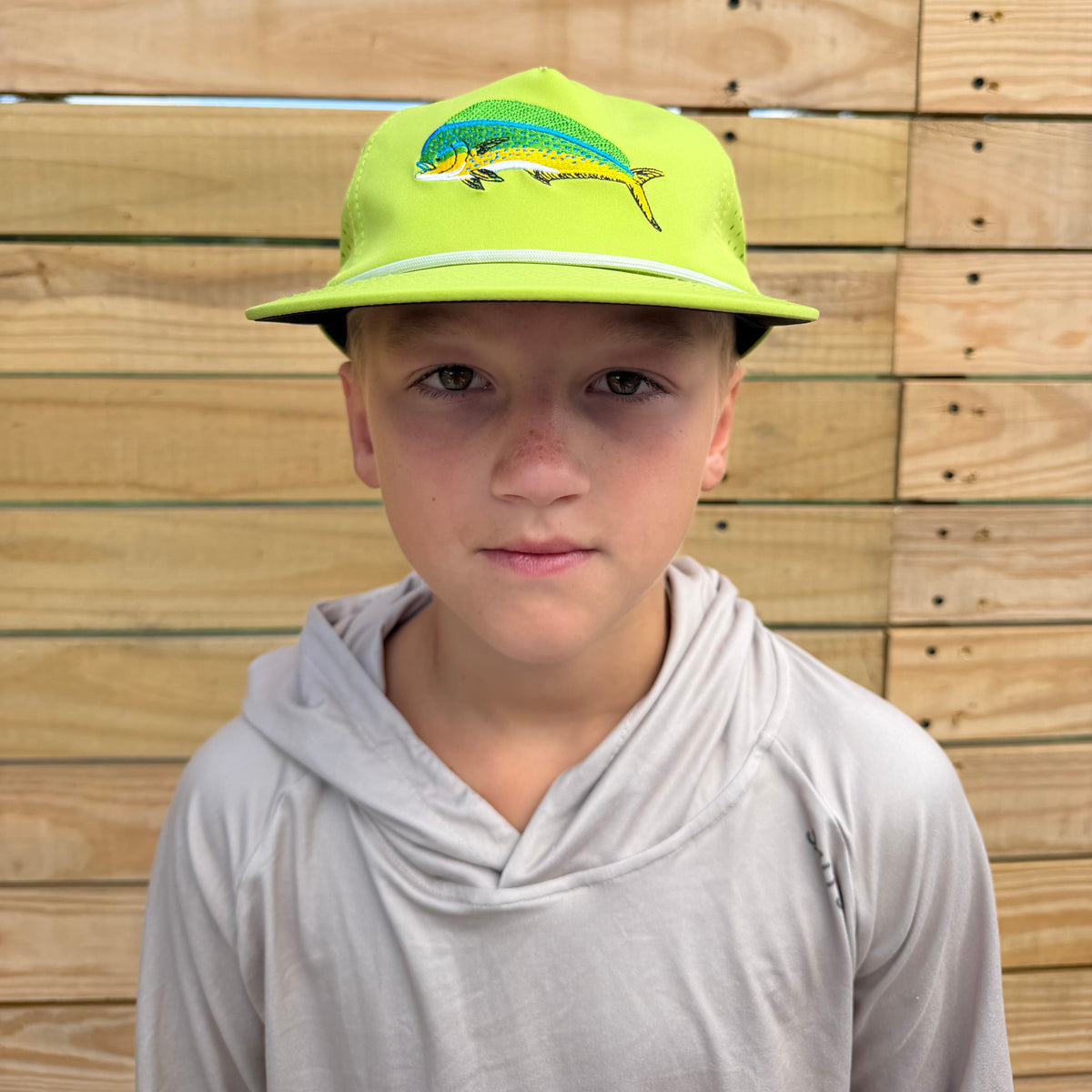 A child wearing a Mahi Mahi Salt SQD baseball cap and a tan hooded sweatshirt, standing against a wooden plank background.
