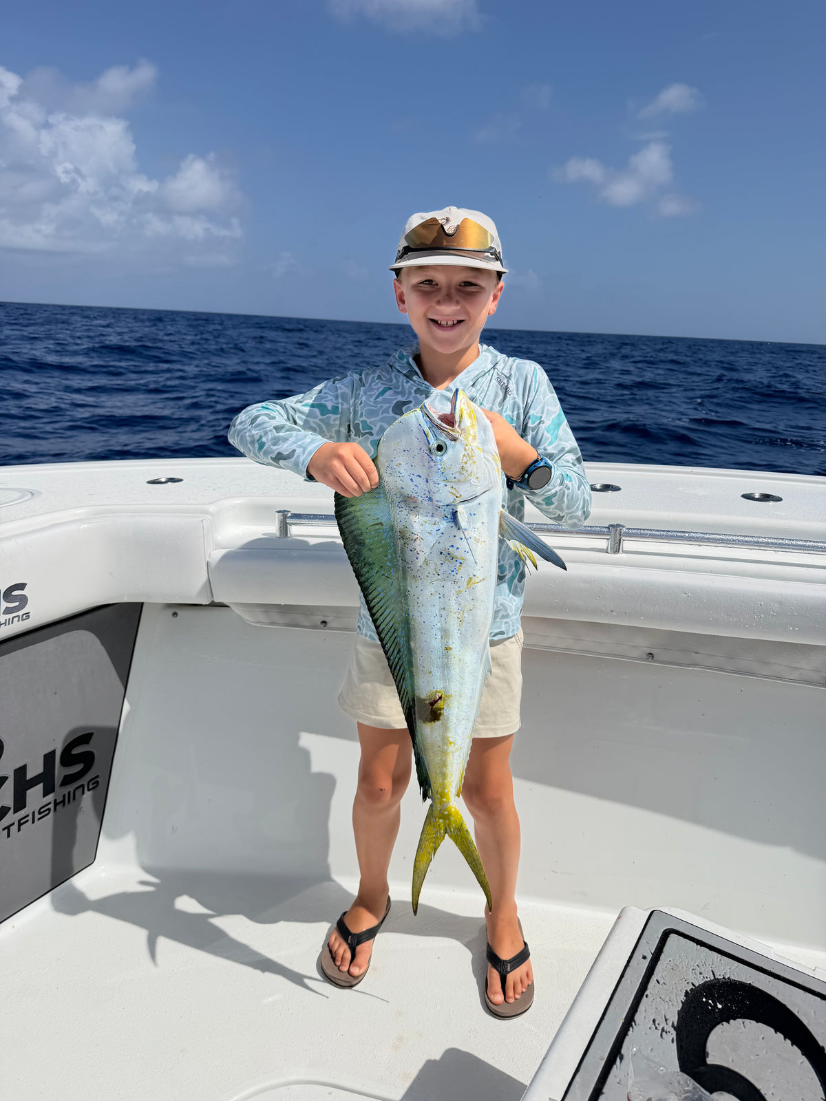 Child standing on a boat deck and holding a large speckled fish with a yellow tail over the ocean