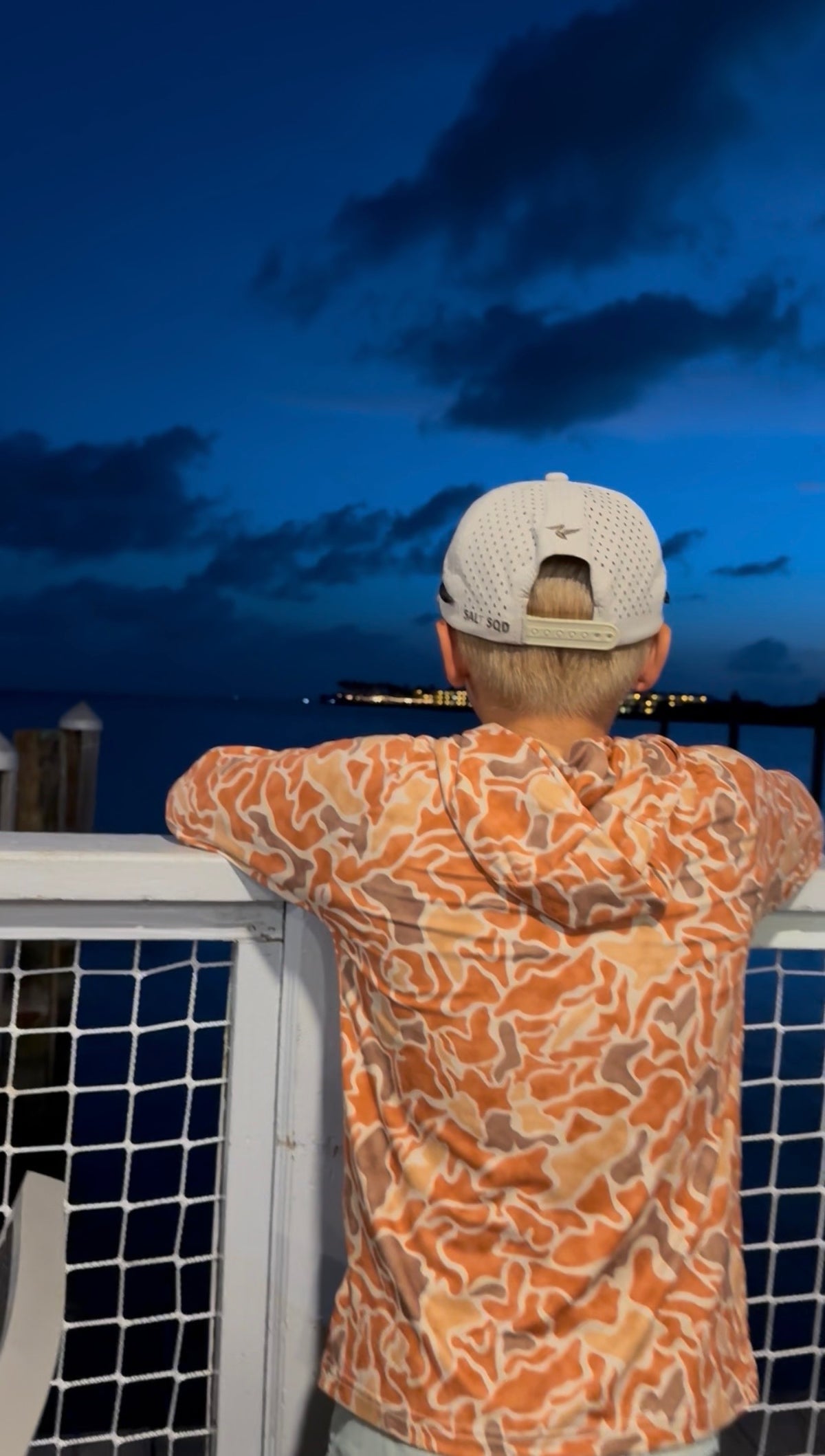 Person in an orange and white patterned shirt and beige mesh cap leaning on a boat railing at dusk