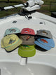 Six embroidered fishing caps in gray, red, brown, blue, yellow, and green arranged on a boat deck