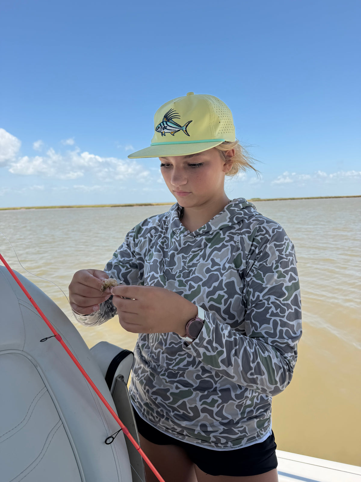 Young person in a camouflage shirt and fish cap adjusting a red line beside a small boat on calm water