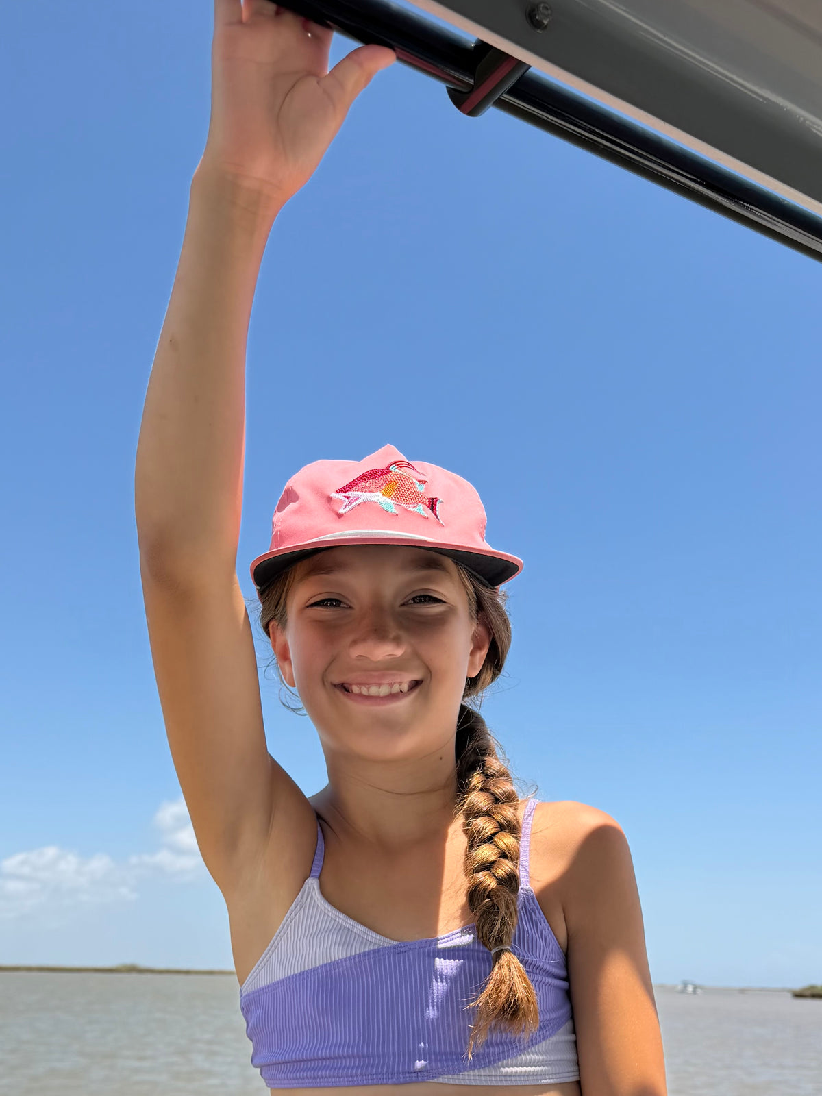 Smiling girl in a pink cap and purple striped top reaching up beneath a metal rail against a blue sky