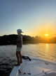 Person standing barefoot on a white boat deck beside rippling water at sunset, wearing a patterned shirt and cap