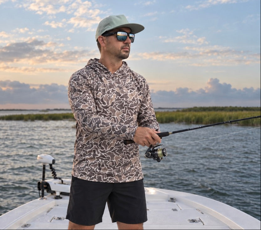 Angler wearing a camouflage hooded shirt and cap, holding a spinning rod on a boat over calm coastal water